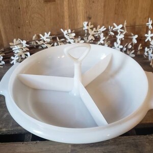 May include: White ceramic serving dish with three divided sections and a handle in the center. The dish is on a wooden surface.