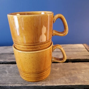 May include: Two brown ceramic mugs stacked on top of each other. The mugs have a speckled glaze and a rounded shape.
