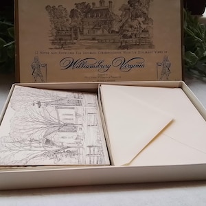 May include: A stationery set featuring a box with the text "Williamsburg Virginia." The box contains notecards and envelopes with illustrations of buildings and trees. The box is beige with a brown lid.