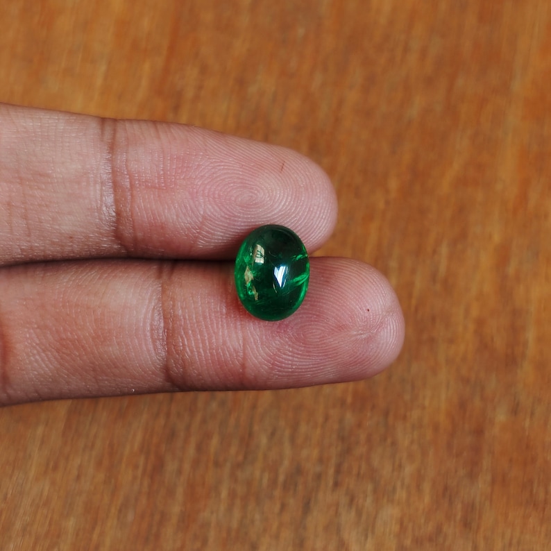 May include: A vibrant green oval-shaped gemstone, likely an emerald, is held between two fingers. The gemstone is smooth and polished, reflecting light.