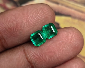 May include: Two emerald green gemstones, cut into square shapes, are held in a hand. The gemstones are faceted and have a brilliant shine.