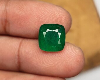 May include: A vibrant, emerald-green gemstone with a cushion cut. The gem has a square shape with rounded corners and a faceted surface that reflects light. The stone is held between fingers, showcasing its color and clarity.