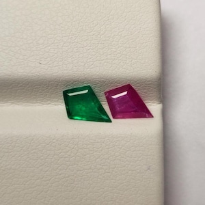 May include: Two gemstones: one emerald green and one magenta. Both are cut in a shield shape with a flat top and angled sides. The gems are displayed on a textured white surface, with a white background.