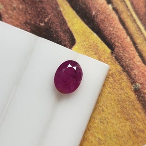 Könnte beinhalten: Ein facettierter, oval geformter Rubin-Edelstein. Der Edelstein hat eine tiefe, satte rote Farbe und liegt auf einer weißen Oberfläche. Der Hintergrund ist eine Mischung aus Gelb, Orange und Braun.