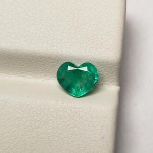 May include: A heart-shaped, emerald-green gemstone. The gem has a faceted surface and a bright, reflective quality. The stone is set against a white background.