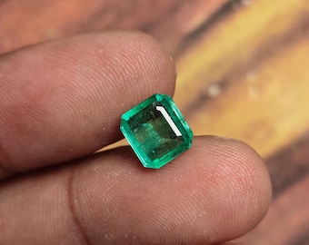 Puede incluir: Una gema de esmeralda verde vibrante, cortada en forma cuadrada con bordes biselados. La gema está engastada en un engaste claro y facetado.