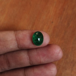May include: A vibrant green oval-shaped gemstone, likely an emerald, is held between two fingers.