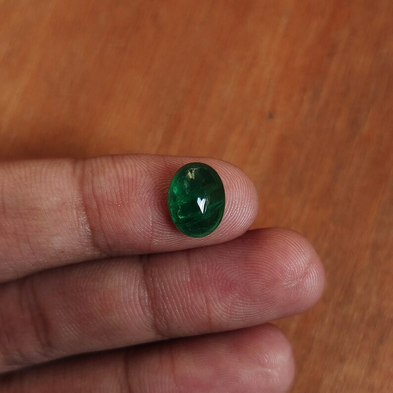 May include: A vibrant green oval-shaped gemstone, likely an emerald, is held between two fingers.