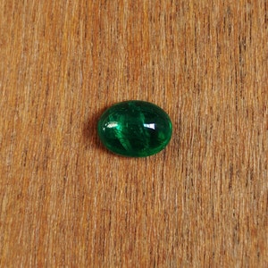 May include: A green oval-shaped gemstone with a smooth, polished surface. The gemstone has a dark green color and is set against a light brown wooden background.
