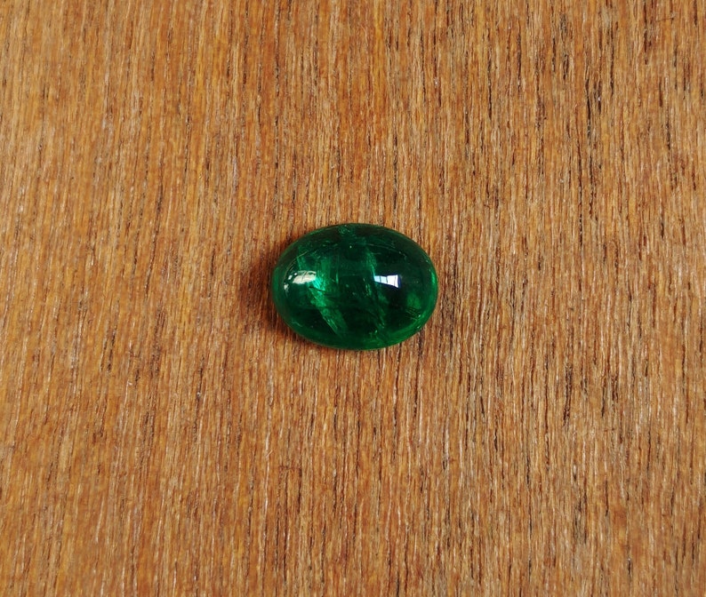 May include: A green oval-shaped gemstone with a smooth, polished surface. The gemstone has a dark green color and is set against a light brown wooden background.