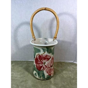May include: A ceramic vase with a cylindrical body and a rounded rim, featuring a hand-painted floral pattern with large red flowers and green leaves on a white background. The vase includes a tall, arched, light brown bamboo handle.