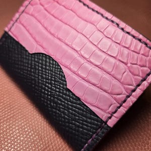 Puede incluir: Un tarjetero de cuero rosa y negro. La mitad superior es rosa con textura de cocodrilo, y la mitad inferior es negra con un patr&oacute;n de trama cruzada. Los bordes est&aacute;n cosidos con hilo negro.