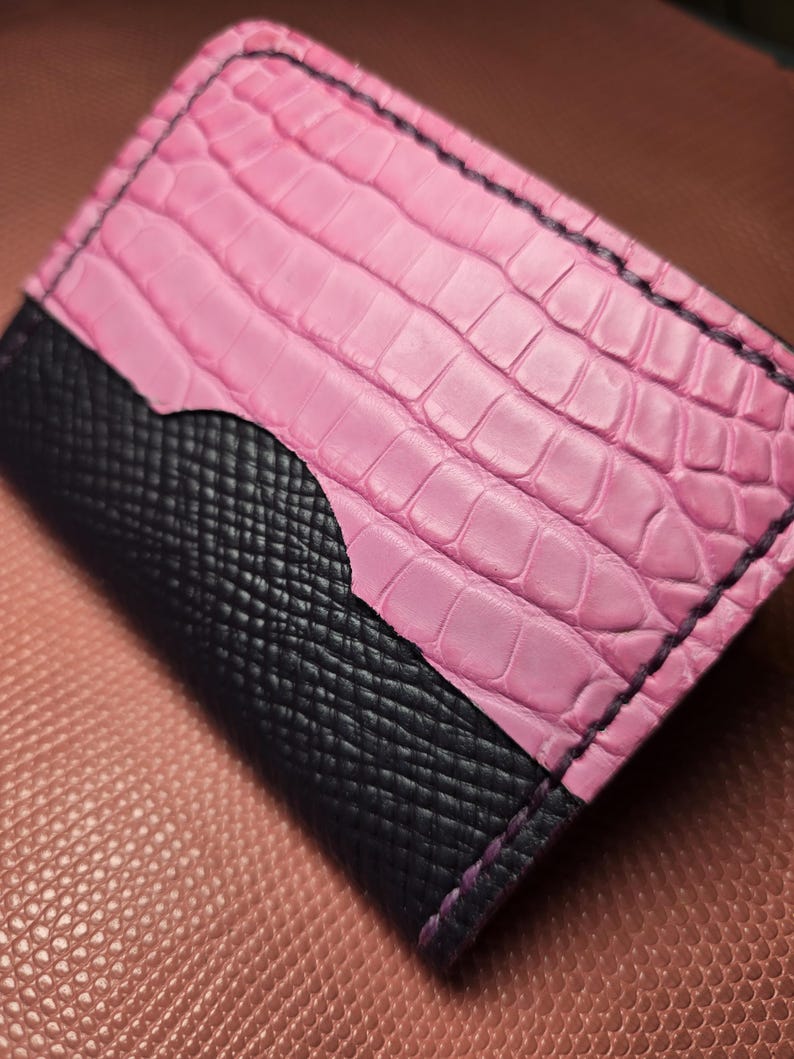 Puede incluir: Un tarjetero de cuero rosa y negro. La mitad superior es rosa con textura de cocodrilo, y la mitad inferior es negra con un patr&oacute;n de trama cruzada. Los bordes est&aacute;n cosidos con hilo negro.