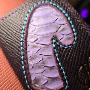 Puede incluir: Primer plano de una cartera de cuero morado oscuro con un dise&ntilde;o escamoso morado claro en el centro. El dise&ntilde;o est&aacute; delineado con costuras turquesas y moradas. La cartera tiene un patr&oacute;n texturizado a cuadros.