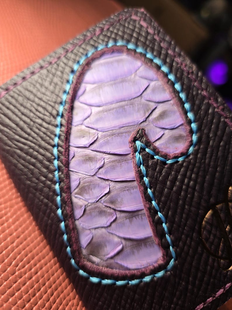 Puede incluir: Primer plano de una cartera de cuero morado oscuro con un dise&ntilde;o escamoso morado claro en el centro. El dise&ntilde;o est&aacute; delineado con costuras turquesas y moradas. La cartera tiene un patr&oacute;n texturizado a cuadros.