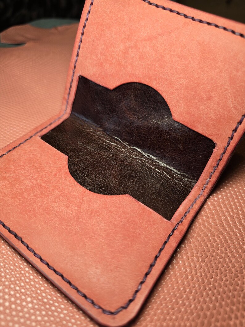 Puede incluir: Cartera de cuero color coral abierta con interior marr&oacute;n oscuro. La cartera presenta un recorte curvo y est&aacute; cosida con hilo morado oscuro. El fondo muestra una superficie texturizada de color coral.