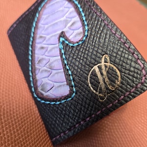 May include: A black leather wallet with a light purple, snake-skin-textured letter "J" stitched with blue thread. The wallet has a gold emblem and purple stitching around the edges.