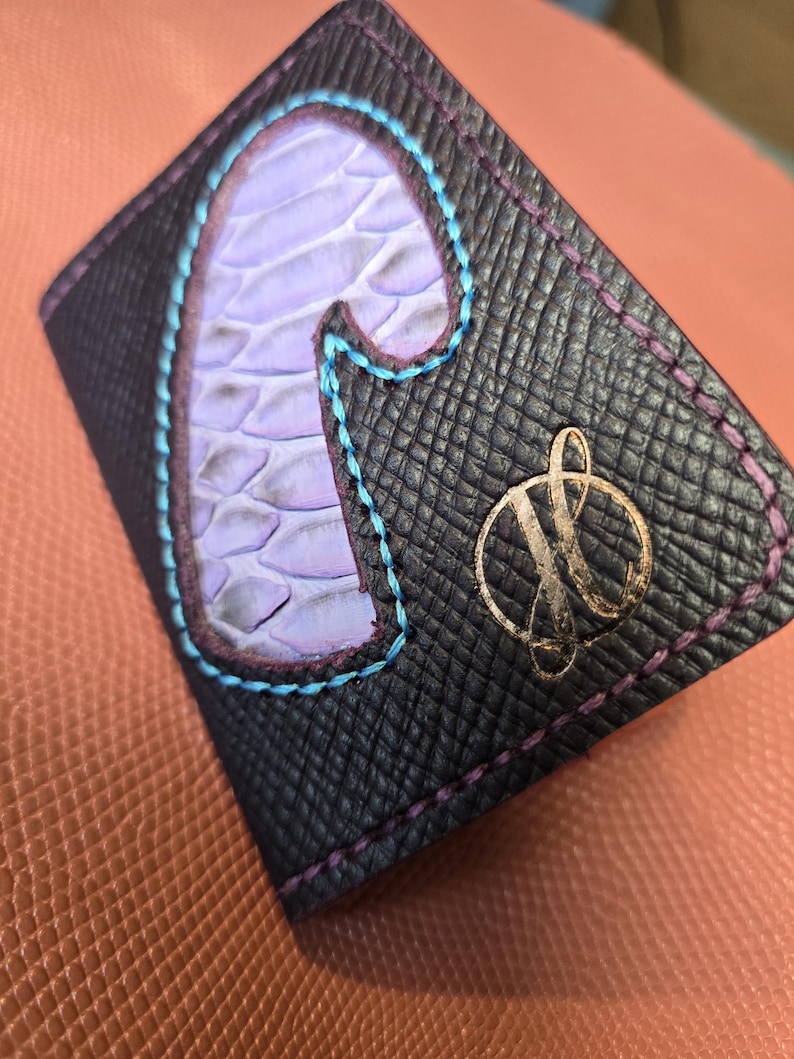 Puede incluir: Cartera de cuero negro con una letra "J" de color p&uacute;rpura claro, con textura de piel de serpiente, cosida con hilo azul. La cartera tiene un emblema dorado y costuras p&uacute;rpuras en los bordes.