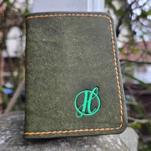 Puede incluir: Cartera de cuero verde oscuro con borde cosido y logotipo de la letra "H" bordada en verde. La cartera tiene un aspecto desgastado y texturizado y costuras de color naranja-marrón.