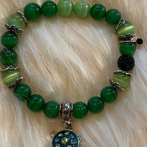 Puede incluir: Un brazalete de piedras verdes con un dije de tortuga de plata. El brazalete está hecho de cuentas redondas con un degradado de color verde claro a verde oscuro. El dije de tortuga tiene diamantes de imitación azules y verdes.