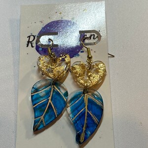Puede incluir: Un par de pendientes de hoja azul y dorada con un dije en forma de corazón. Los pendientes están hechos de resina y tienen un gancho chapado en oro.