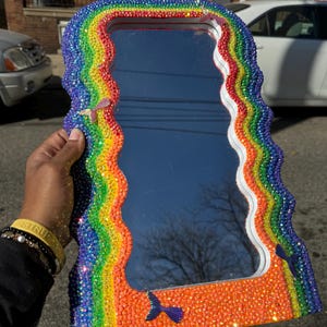 Pode incluir: Um espelho arco-íris com uma moldura ondulada adornada com pedras preciosas. O espelho é decorado com pequenas pedras preciosas iridescentes e apresenta um design de cauda de sereia na parte inferior.