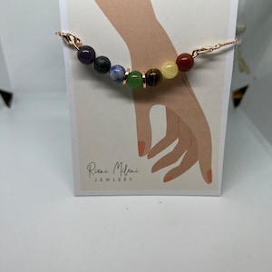Puede incluir: Un brazalete de cadena de oro rosa con siete cuentas de piedras preciosas de colores. Las cuentas son de amatista, obsidiana negra, lapislázuli, aventurina verde, ojo de tigre, citrino y jaspe rojo. El brazalete está sobre una tarjeta blanca con el texto "Riani Milani Jewelry".
