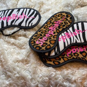 Puede incluir: Cuatro máscaras para dormir con una banda elástica negra. Dos máscaras son de estampado de cebra en blanco y negro con el texto "Sleep Bish..." en rosa. Dos máscaras son de estampado de leopardo marrón y negro con el texto "Sleep Bish..." en rosa.