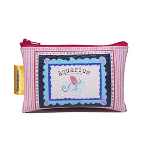 May include: A rectangular coin purse with a pink and white striped pattern. The purse features a framed design with the word "Aquarius" and a zodiac symbol. It has a red zipper and a yellow tag.
