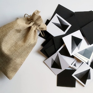 Può includere: Un sacco di juta con chiusura a coulisse si trova accanto a un mucchio di carte bianche e nere con un design geometrico. Le carte presentano un triangolo nero con un motivo sfumato.