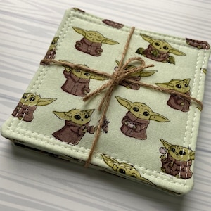 Puede incluir: Conjunto de cuatro posavasos de tela verde con un borde blanco cosido. Los posavasos presentan un patr&oacute;n repetido de un personaje de dibujos animados con orejas grandes y una t&uacute;nica marr&oacute;n. Los posavasos est&aacute;n atados juntos con una cuerda.