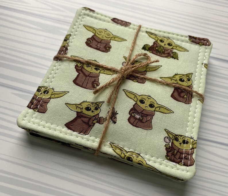 Puede incluir: Conjunto de cuatro posavasos de tela verde con un borde blanco cosido. Los posavasos presentan un patr&oacute;n repetido de un personaje de dibujos animados con orejas grandes y una t&uacute;nica marr&oacute;n. Los posavasos est&aacute;n atados juntos con una cuerda.