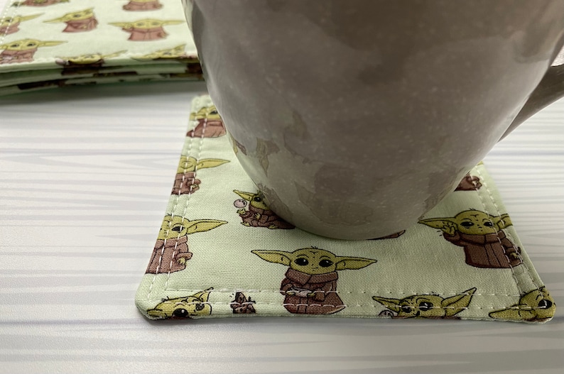 Puede incluir: Un posavasos cuadrado verde con fondo blanco y un patr&oacute;n repetido de personajes de dibujos animados. El posavasos tiene un borde cosido y est&aacute; sobre una superficie blanca con una taza encima.