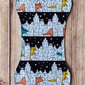 May include: A black and white fabric with a repeating pattern of cityscapes and dinosaurs. The dinosaurs are orange, yellow, and green. The cityscapes are white and black.