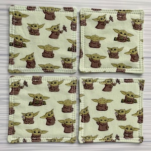 Puede incluir: Conjunto de cuatro posavasos cuadrados de tela con un patr&oacute;n repetido de un personaje alien&iacute;gena verde con orejas grandes. Los posavasos son beige con un borde blanco cosido.