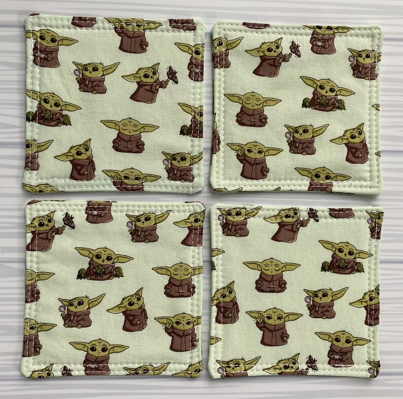 Puede incluir: Conjunto de cuatro posavasos cuadrados de tela con un patr&oacute;n repetido de un personaje alien&iacute;gena verde con orejas grandes. Los posavasos son beige con un borde blanco cosido.