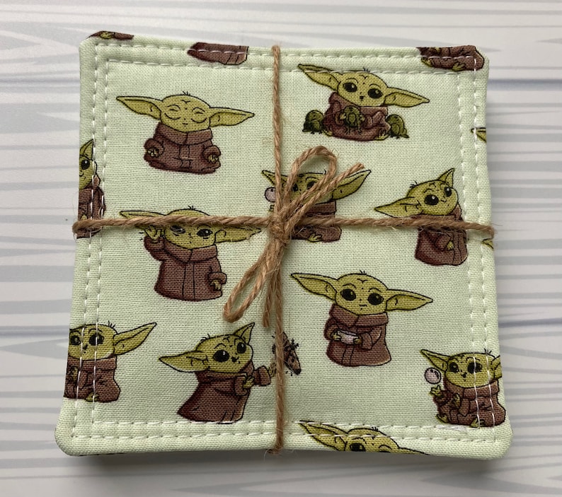 Puede incluir: Conjunto de cuatro posavasos de tela verde con un patr&oacute;n repetido de un personaje de dibujos animados de la franquicia Star Wars. Los posavasos est&aacute;n atados con cuerda.