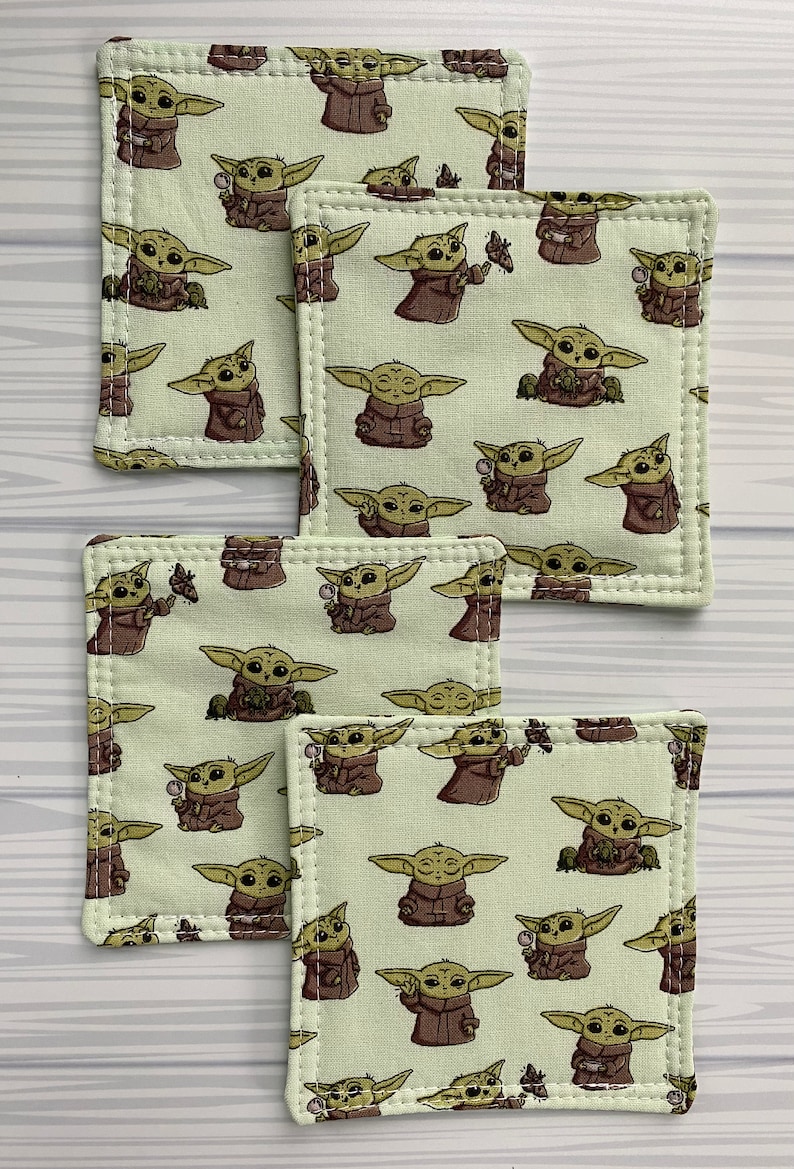 Puede incluir: Conjunto de cinco posavasos de tela verde con un patr&oacute;n repetido de un personaje de dibujos animados de la franquicia Star Wars. El personaje es verde y marr&oacute;n y sostiene un objeto peque&ntilde;o en su mano.