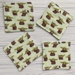 Puede incluir: Cuatro posavasos cuadrados de tela verde con un patr&oacute;n repetido de un personaje de dibujos animados con ojos grandes y orejas puntiagudas. El personaje lleva una t&uacute;nica marr&oacute;n y sostiene un objeto peque&ntilde;o en su mano.