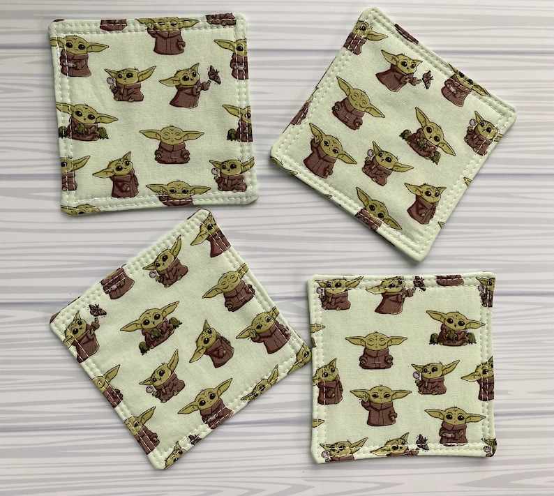 Puede incluir: Cuatro posavasos cuadrados de tela verde con un patr&oacute;n repetido de un personaje de dibujos animados con ojos grandes y orejas puntiagudas. El personaje lleva una t&uacute;nica marr&oacute;n y sostiene un objeto peque&ntilde;o en su mano.