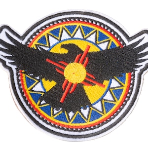 Può includere: Toppa ricamata con un'aquila nera con le ali spiegate, centrata su un disegno circolare. Il disegno include un simbolo solare giallo con raggi rossi, circondato da motivi geometrici blu e gialli e un bordo bianco.