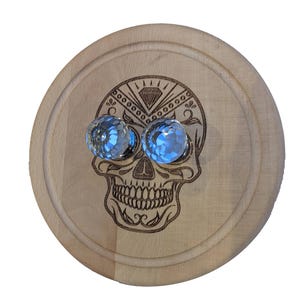 Puede incluir: Una tabla de cortar redonda de madera con un diseño de calavera grabado con láser. Dos esferas de vidrio azul facetadas sirven como ojos. La calavera tiene detalles decorativos y una forma de diamante encima de los ojos.