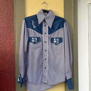 May include: A vintage blue and white gingham shirt with long sleeves. The shirt features a dark blue yoke and cuffs with embroidered figures. White piping accents the collar, pockets, and button placket. The shirt is hanging on a metal hanger.