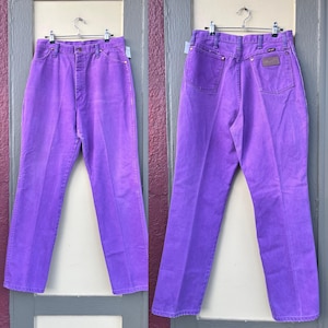 May include: A pair of vibrant purple Wrangler jeans, displayed front and back. The jeans are hung on a metal hanger, showcasing the classic five-pocket design, a button and zipper closure, and a leather patch on the back. The jeans are made of denim.