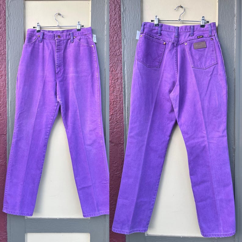 May include: A pair of vibrant purple Wrangler jeans, displayed front and back. The jeans are hung on a metal hanger, showcasing the classic five-pocket design, a button and zipper closure, and a leather patch on the back. The jeans are made of denim.