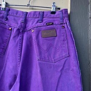 May include: Close-up of purple Wrangler jeans hanging on a metal hanger. The jeans have a brown leather patch with the Wrangler logo on the back pocket. The jeans have a classic five-pocket design and are made of a durable denim material.