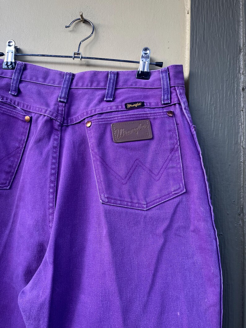 May include: Close-up of purple Wrangler jeans hanging on a metal hanger. The jeans have a brown leather patch with the Wrangler logo on the back pocket. The jeans have a classic five-pocket design and are made of a durable denim material.