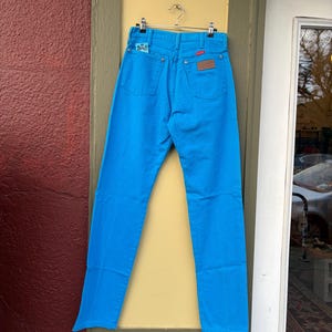 May include: Bright blue denim jeans hanging on a metal hanger. The jeans have a classic five-pocket design with a brown leather patch and a small embroidered detail on the back pocket. The jeans are a vibrant, solid color.