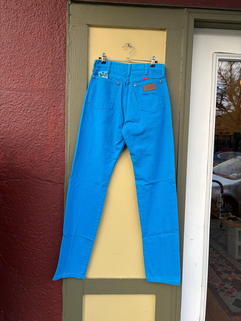 May include: Bright blue denim jeans hanging on a metal hanger. The jeans have a classic five-pocket design with a brown leather patch and a small embroidered detail on the back pocket. The jeans are a vibrant, solid color.