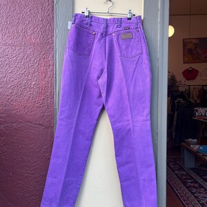 May include: Bright purple Wrangler jeans hanging on a metal hanger. The jeans have a classic five-pocket design with a brown leather patch on the back pocket. The jeans are displayed in a doorway with a purple wall on the left.